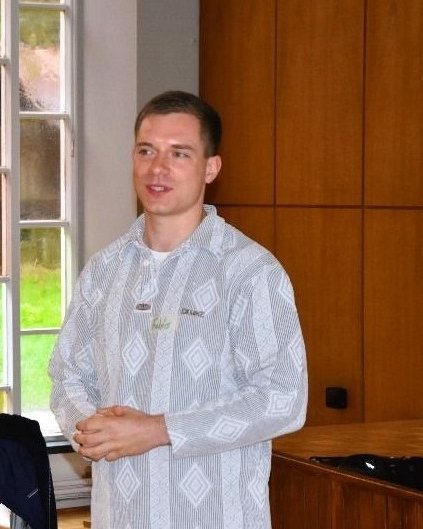 Fabio Banet steht in einem Seminarraum mit Holzwand und hohen Fenstern und spricht zu einer Gruppe. Er trägt ein helles, gemustertes Hemd und ein Namensschild.