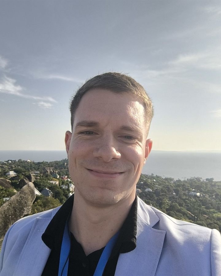 Fabio Banet lächelt in die Kamera. Er trägt ein hellblaues Sakko, hinter ihm liegen eine grüne Hügellandschaft mit Häusern und eine weite Wasserfläche.